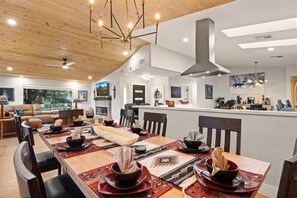 A modern dining area with a long wooden table set for a meal, a kitchen in the background, and a view of the outdoors. - Gorgeous Designer Home, Sedona Red Rock Basin Views, Outdoor Living Entertainment Patio w/ Firepit & BBQ! The Den - S079 (Sedona)