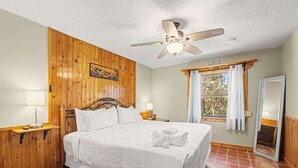 A bedroom with a wooden wall, a bed with white bedding, a ceiling fan, a window with curtains, and a mirror. - Aqua Queen - Pool 3BR (St. Augustine)