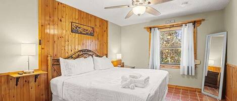 Un dormitorio con pared de madera, cama con ropa blanca, ventilador de techo, ventana con cortinas y un espejo.