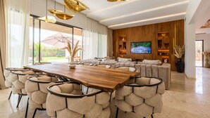 Ein großzügiges Wohnzimmer mit einem großen Esstisch aus Holz, umgeben von bequemen, gepolsterten Stühlen. Eine gemütliche Couch und ein Flachbildfernseher stehen an der Wand, darüber befindet sich ein Holzpaneel mit Regalen.
