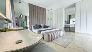 Ein modernes Schlafzimmer mit einem großen, ovalen Waschbecken, einem Bett mit grauer Bettwäsche, einer gepolsterten grauen Bank und einem großen Spiegel.