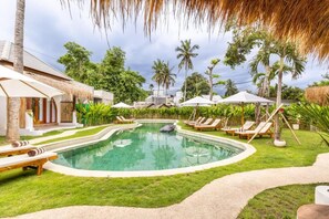 A swimming pool with lounge chairs, a thatched-roof structure, and lush greenery.