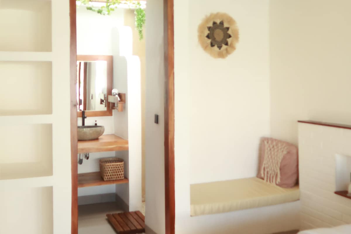 Un dormitorio con cama, cabecera de madera, banqueta con cojines, un adorno en la pared y un acceso a un baño con espejo y lavamanos.