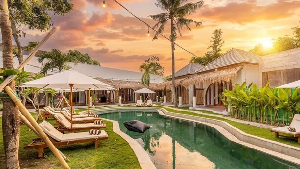 A pool area with lounge chairs and umbrellas, surrounded by thatched-roof buildings and palm trees. - Roh Villas Kuta Lombok (Kuta)