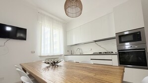 Eine moderne Küche mit weißen Schränken, einem Holztisch, weißen Stühlen, einem Flachbildfernseher, einem eingebauten Backofen und einer Pendelleuchte.