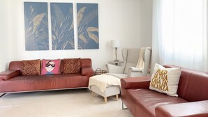 Ein Wohnzimmer mit einem roten Sofa, einem weißen Sessel, einem kleinen Tisch und einer Lampe. Zwei Wandklimaanlagen und ein Fenster mit Vorhängen sind ebenfalls vorhanden.