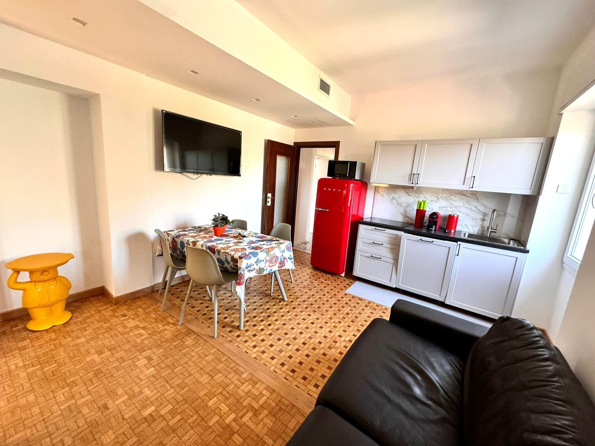 Un espacio habitable compacto con comedor, cocina con armarios blancos, refrigerador rojo y televisor de pantalla plana montado en la pared.