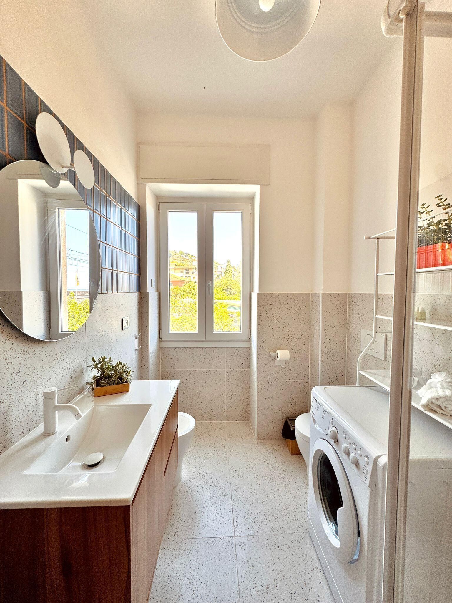 A modern bathroom with a sink, mirror, window, and washing machine.