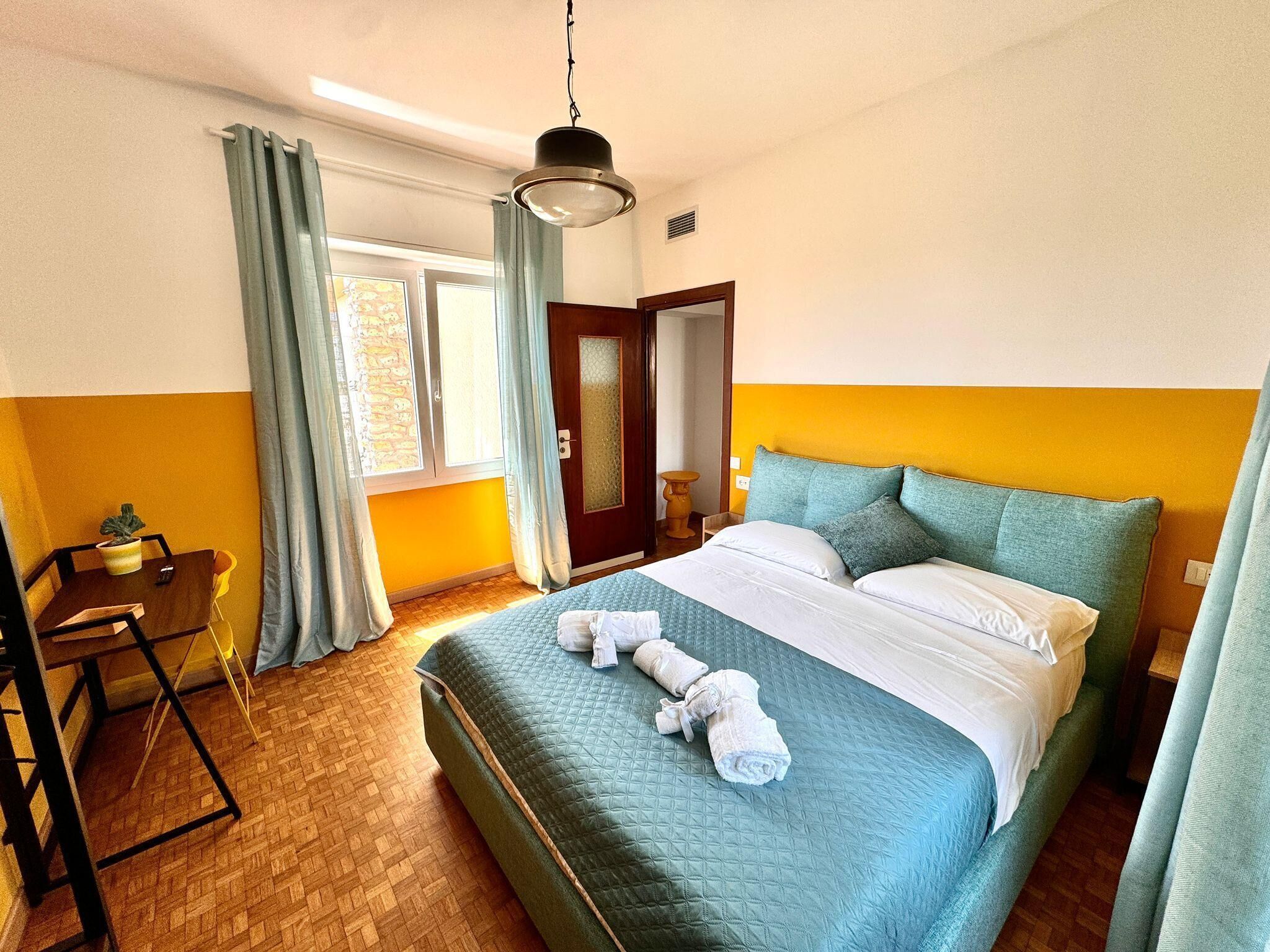 A hotel room with a bed, a desk, a chair, a window with curtains, and a door.