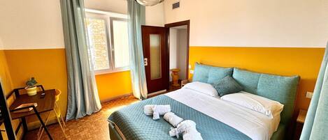 A hotel room with a bed, a desk, a chair, a window with curtains, and a door.