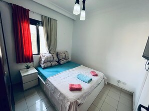 A bedroom with a bed, a nightstand, a window with curtains, and a ceiling light. - The View - Community - 2 Room Apartment (Red Sea Governorate)