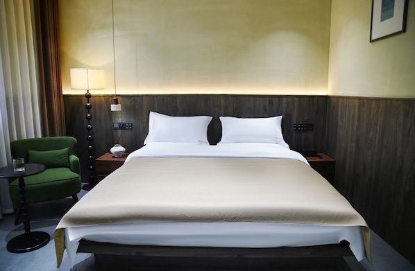 A hotel room with a neatly made bed, a green armchair, a bedside table with a lamp, and a wall-mounted control panel.