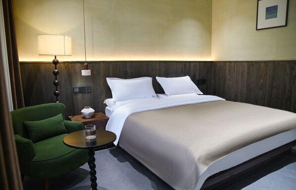 Habitación de hotel con una cama, un sillón verde, una mesita con lámpara y un panel de control en la pared.