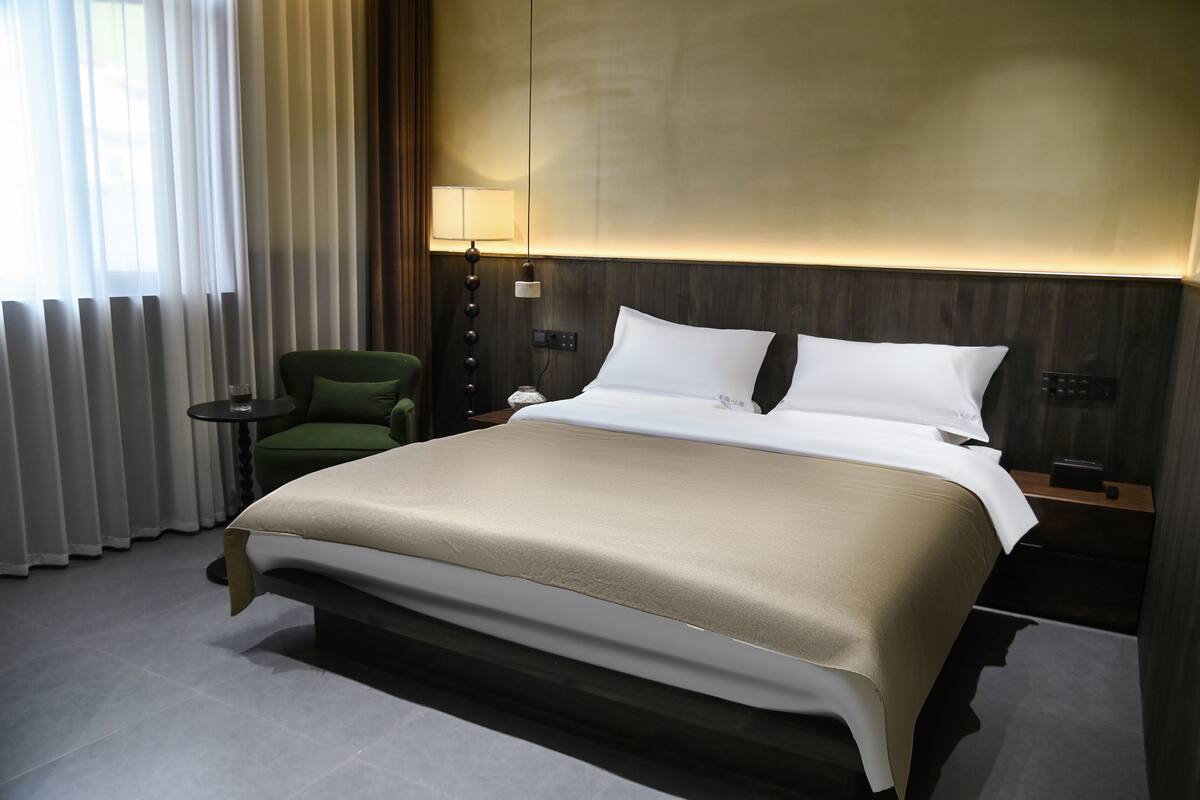 Een hotelkamer met een groot bed, een groene fauteuil, een nachtkastje en een lamp.