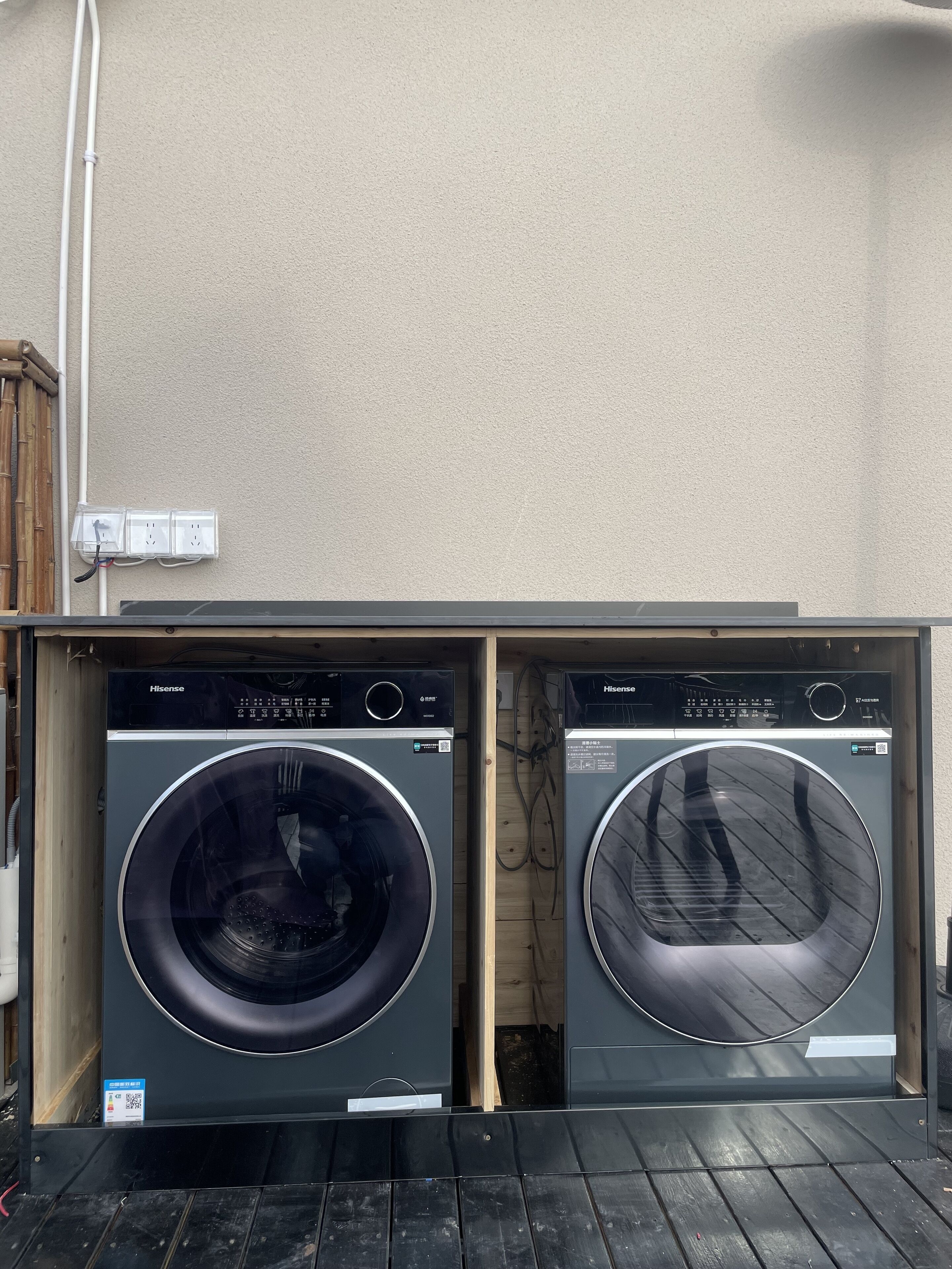 Two Hisense washing machines in a wooden cabinet with a beige wall and a power outlet above.
