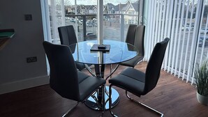 A modern dining area with a round glass table and four black chairs, large windows with white blinds, and a view of a residential street. - Christchurch Apartment, Great Location With Parking (Christchurch)