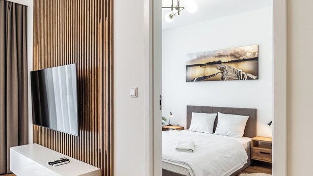 A modern bedroom with a large flat-screen TV mounted on a wall, a bed with white bedding, a nightstand, and a painting of a sunset over a bridge. - RentPlanet - Apartament Przemysłowa (Katowice)
