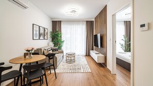 A modern living room with a wooden dining table, black chairs, a sofa, a TV, and a large window with curtains. - RentPlanet - Apartament Przemysłowa (Katowice)
