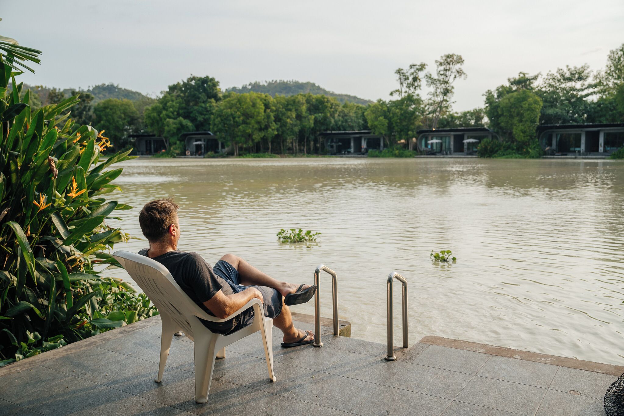 A man sitting on a chair by a water body with a view of a forested area and buildings in the background.