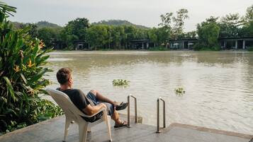 A man sitting on a chair by a water body with a view of a forested area and buildings in the background.