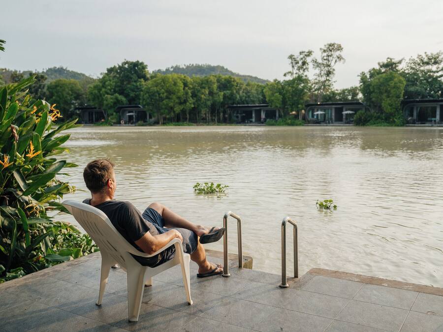 A man sitting on a chair by a water body with a view of a forested area and buildings in the background.