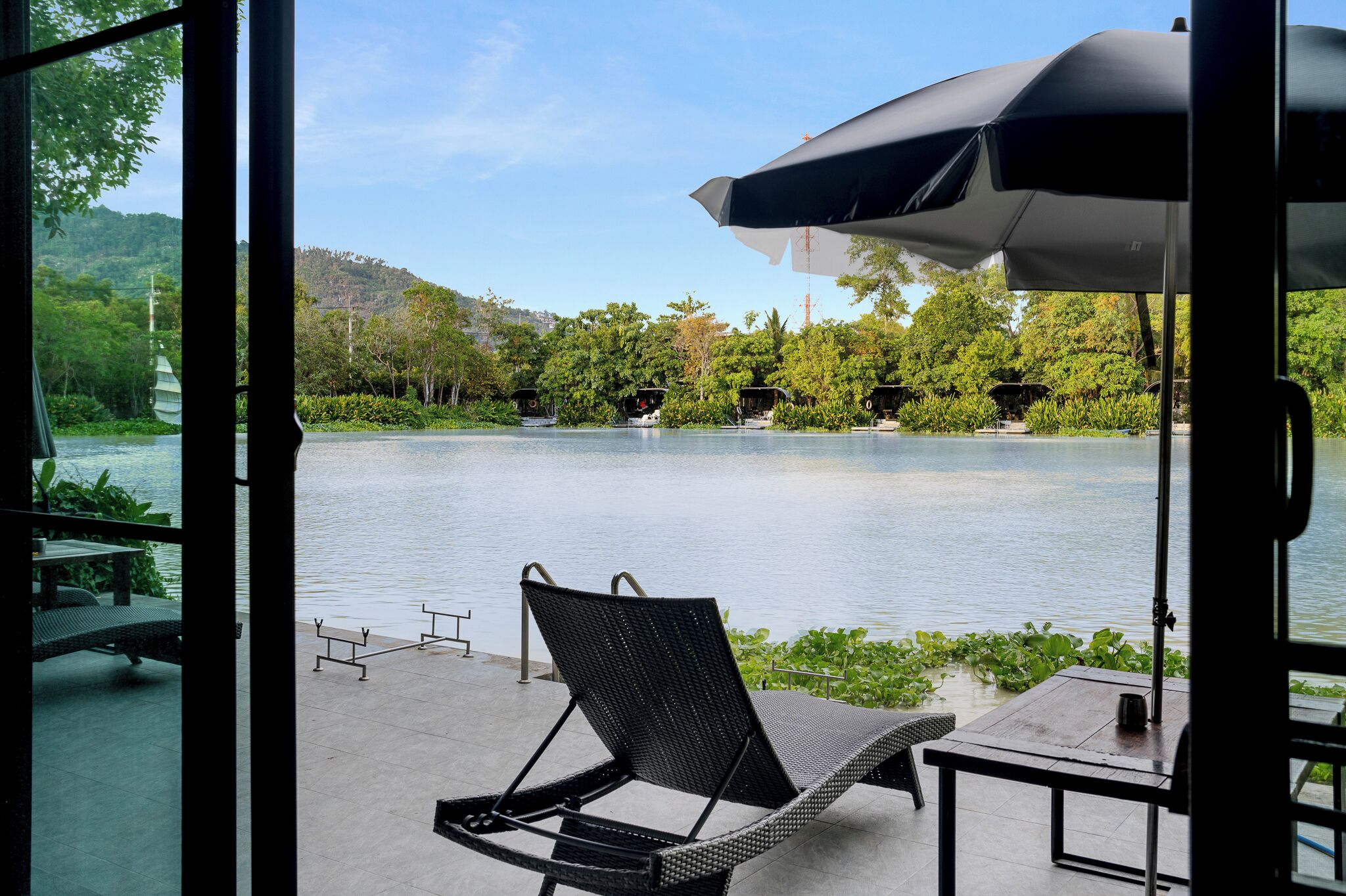 A view from a room with a large window overlooking a lake, a lounge chair, and a table with an umbrella.