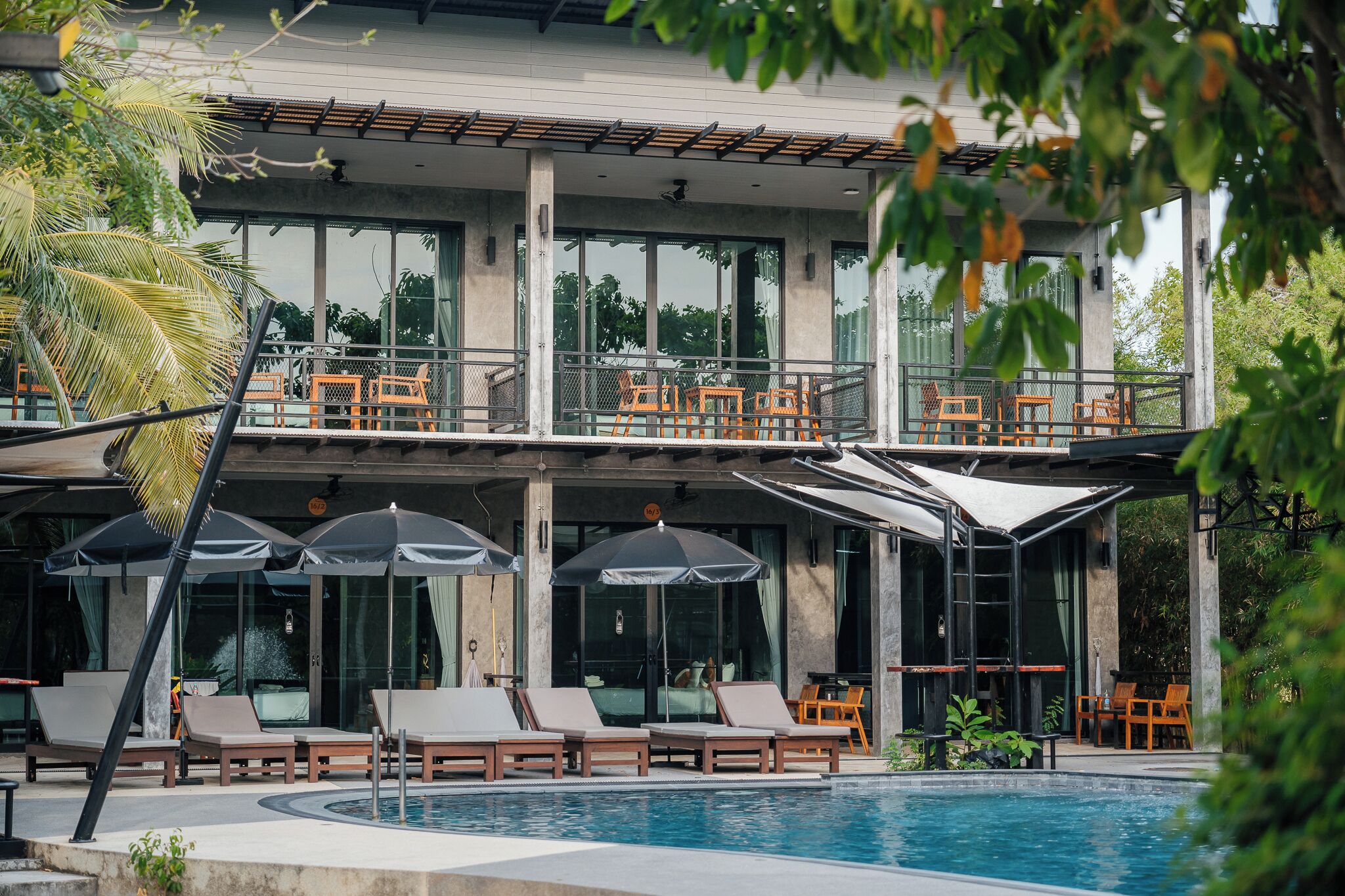 Un espace aménagé au bord de la piscine, avec des chaises longues et des parasols, un espace de détente couvert avec des tables, et une vue sur la piscine.