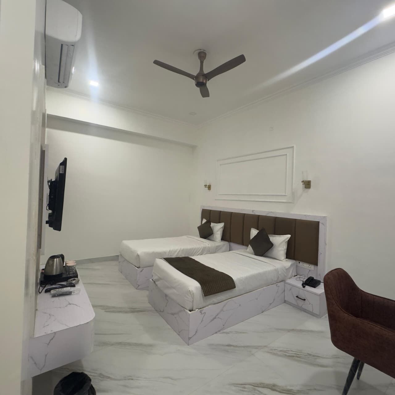 A hotel room with two beds, a ceiling fan, a marble floor, a small desk with a chair, and a TV mounted on the wall.