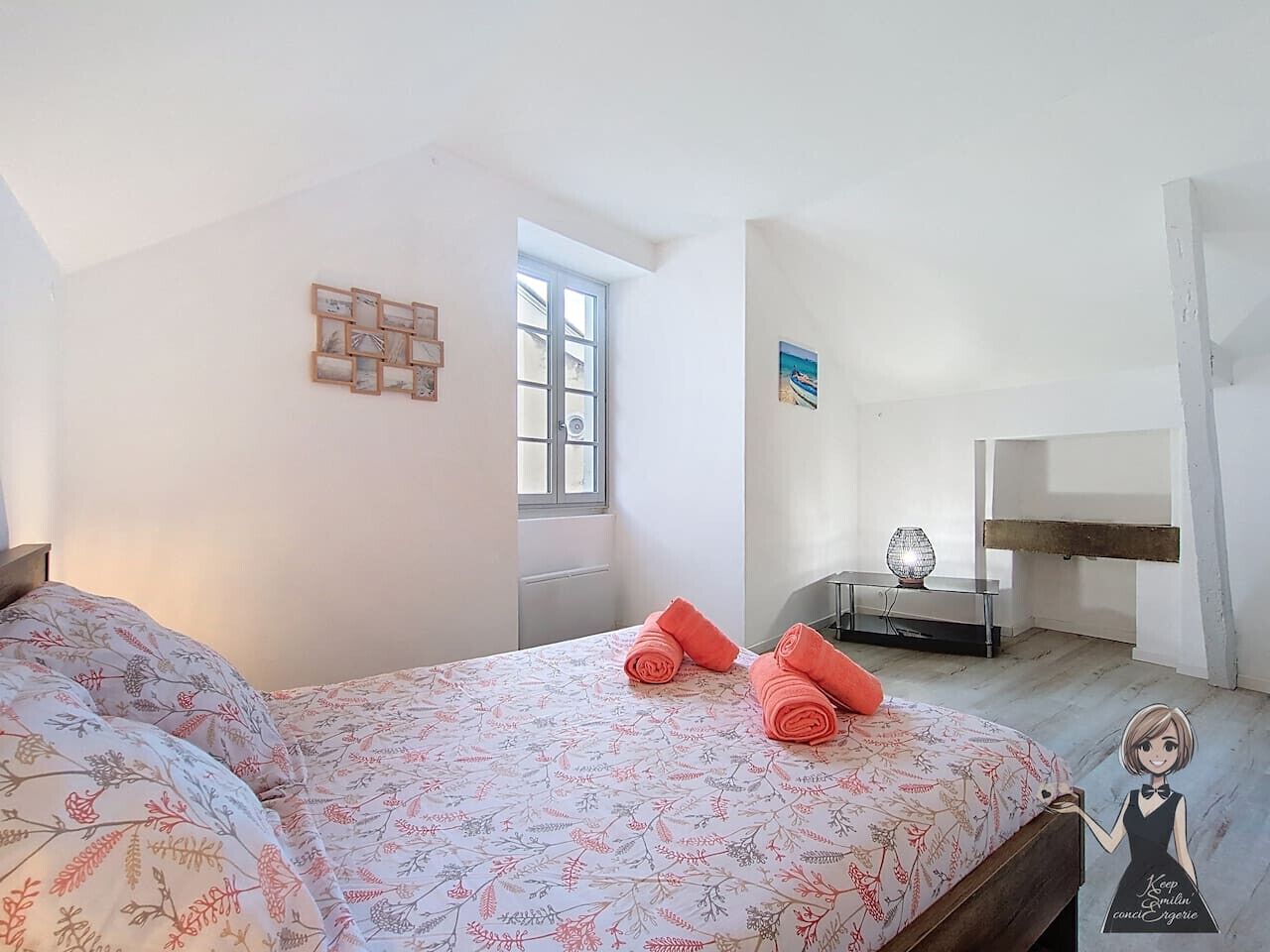 A bedroom with a bed, two rolled towels, a window, a bedside table with a lamp, and a wall-mounted shelf.