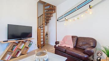 Un salon avec un canapé en cuir marron, un téléviseur à écran plat posé sur un meuble en bois, un escalier et une guirlande lumineuse accrochée au mur.