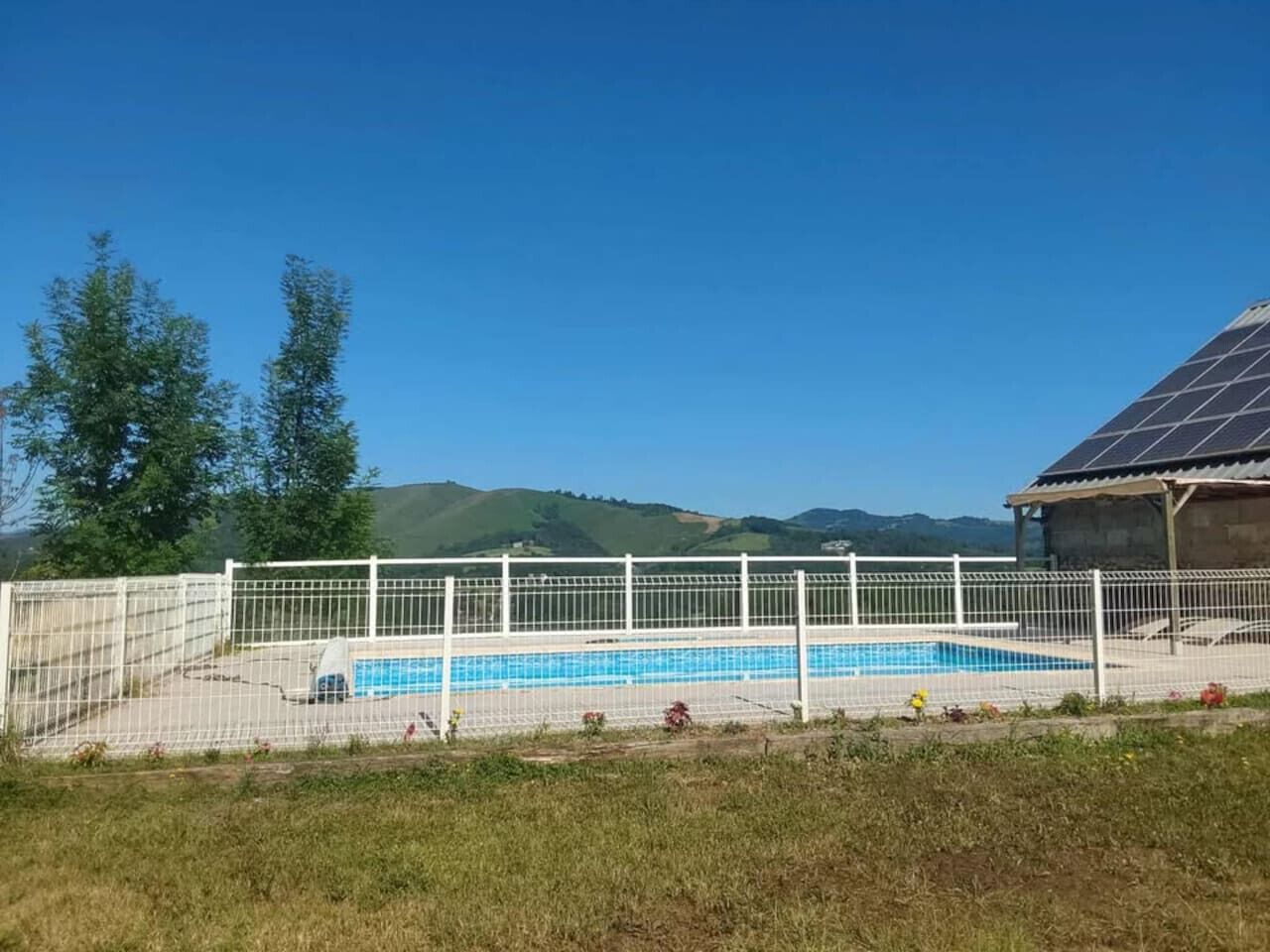 A swimming pool with a white fence, surrounded by grass and trees, with a building with a solar panel roof in the background.