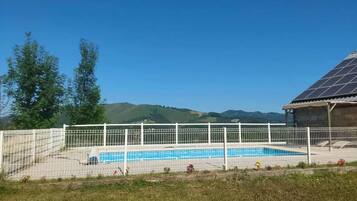 A swimming pool with a white fence, surrounded by grass and trees, with a building with a solar panel roof in the background.