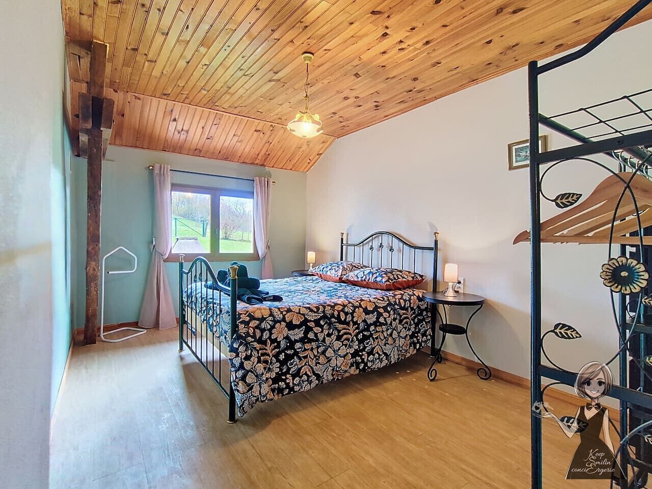 A bedroom with a wooden ceiling, a bed with a floral bedspread, a small table with a lamp, and a window with curtains.