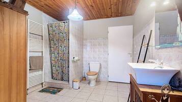Een badkamer met een houten plafond, een wit toilet, een douche met een kleurrijk gordijn en een witte wastafel met een spiegel erboven.