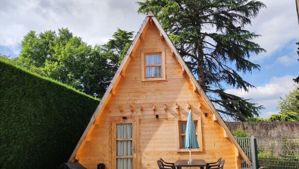 Een houten hut met een puntig dak, een klein terras en een tafel met stoelen.