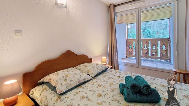 A bedroom with a wooden headboard, floral bedding, a nightstand with a lamp, and a view of a balcony with a railing. - Le Refuge, Cocon pour 8 avec Spa & Piscine chauf. (Cauterets)