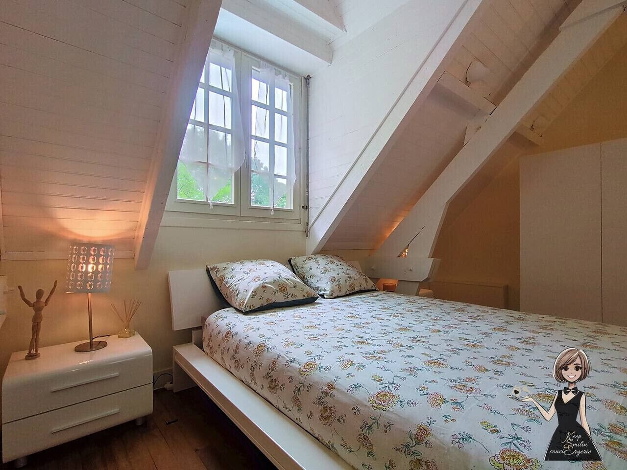 A bedroom with a sloped ceiling, a bed with floral bedding, a bedside lamp, and a window with curtains.