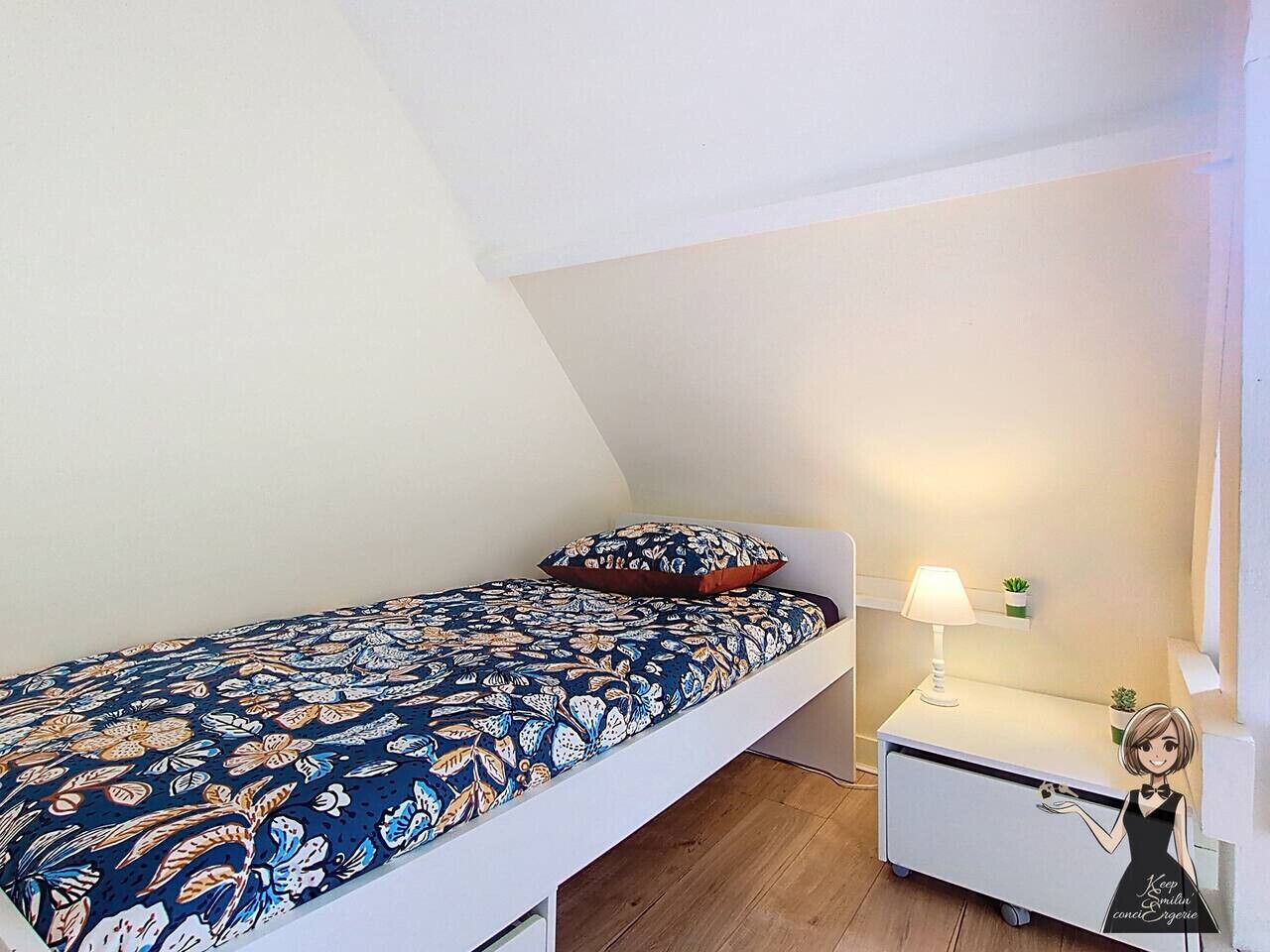 A small, single-bed room with a patterned bedspread, a bedside table with a lamp, and a small potted plant.