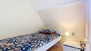 A small, single-bed room with a patterned bedspread, a bedside table with a lamp, and a small potted plant.