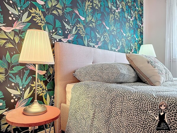 A bedroom with a patterned bedspread, a round side table with a lamp, and a floral wallpaper.
