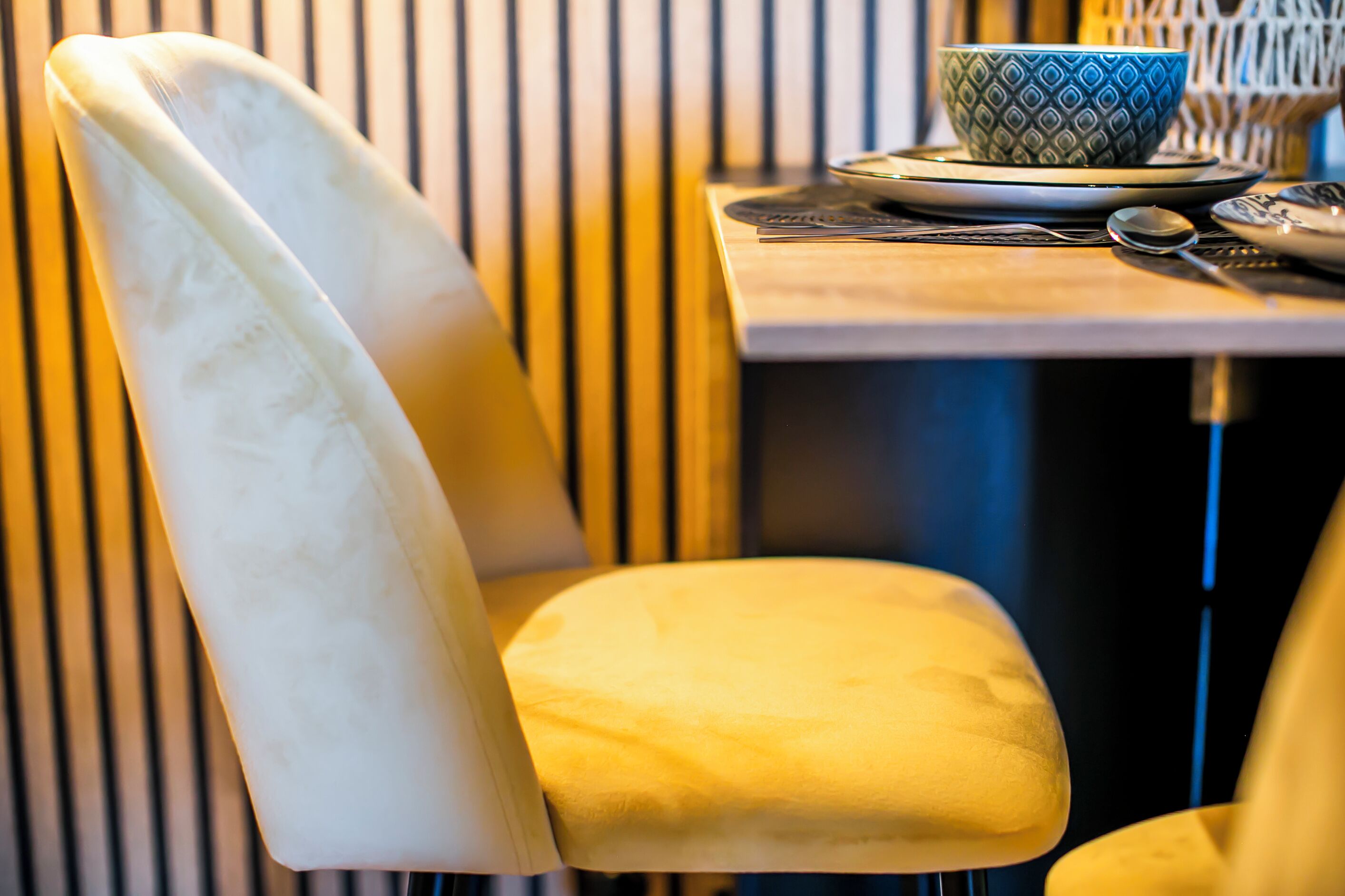 Een stoel met een witte rugleuning en een geel kussen, een tafel met een blauwe kom met patroon en een lepel, en verticale jaloezieën op de achtergrond.