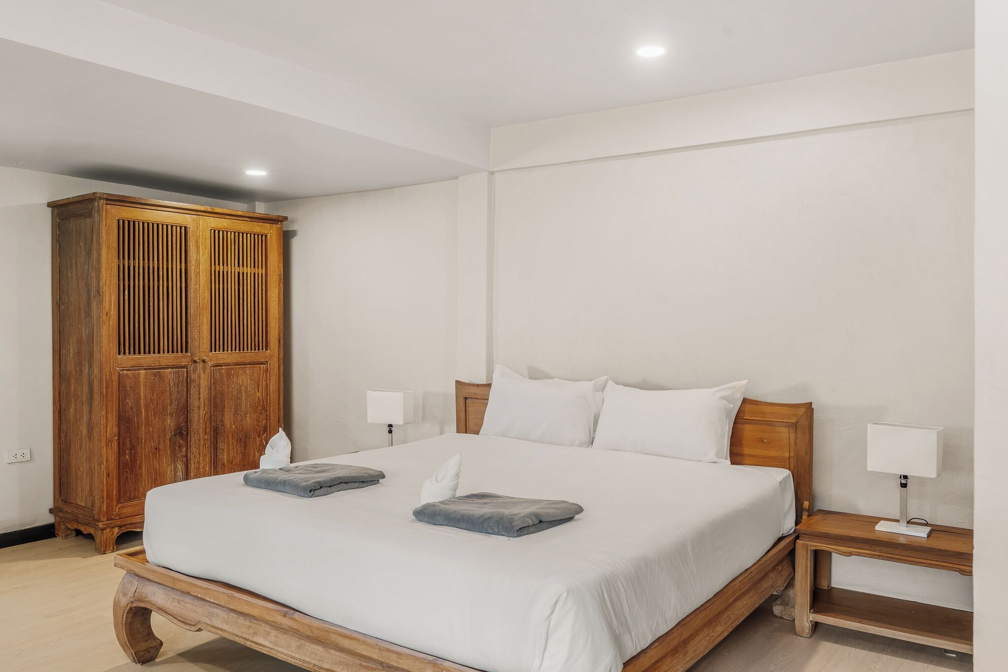 Un dormitorio con estructura de cama de madera, ropa de cama blanca, dos mesitas de noche con lámparas y un armario de madera.