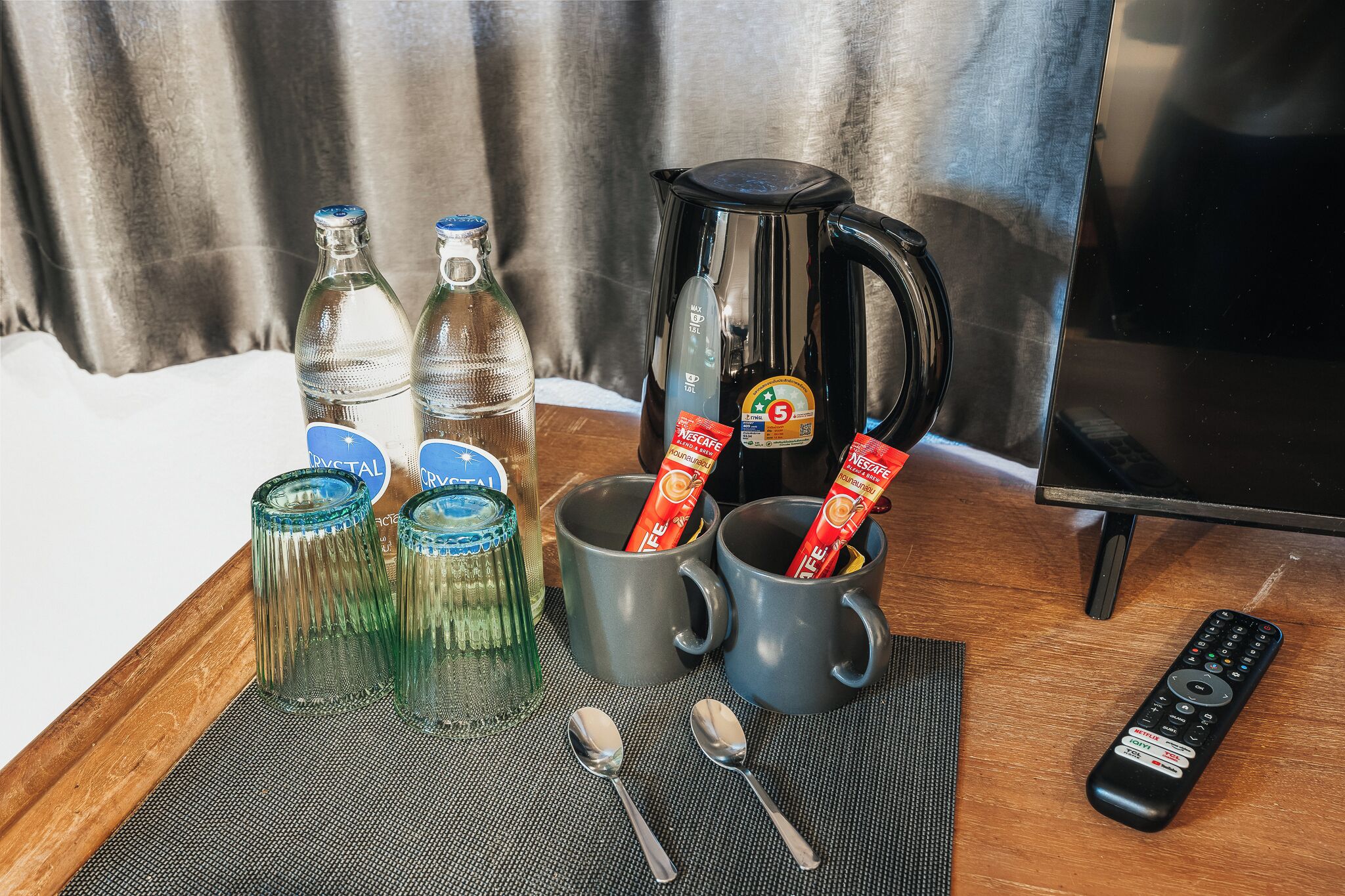 A tray with two clear bottles, two gray mugs with Nescafé packets, a black coffee maker, two spoons, and a remote control.