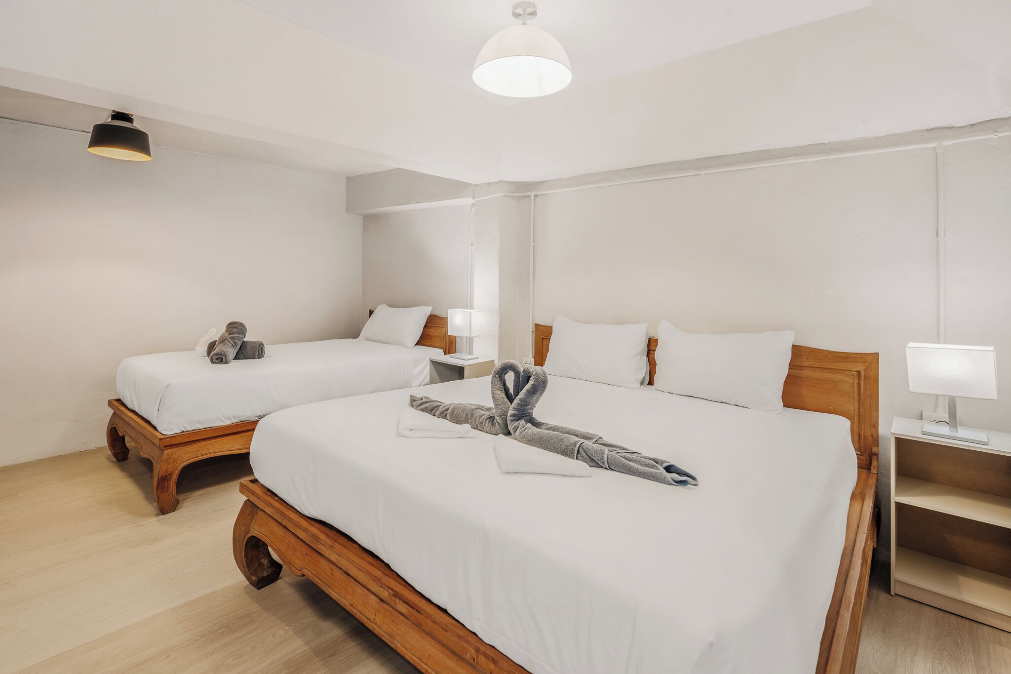 A hotel room with two beds, wooden headboards, white bedding, a bedside table with a lamp, and a ceiling light.