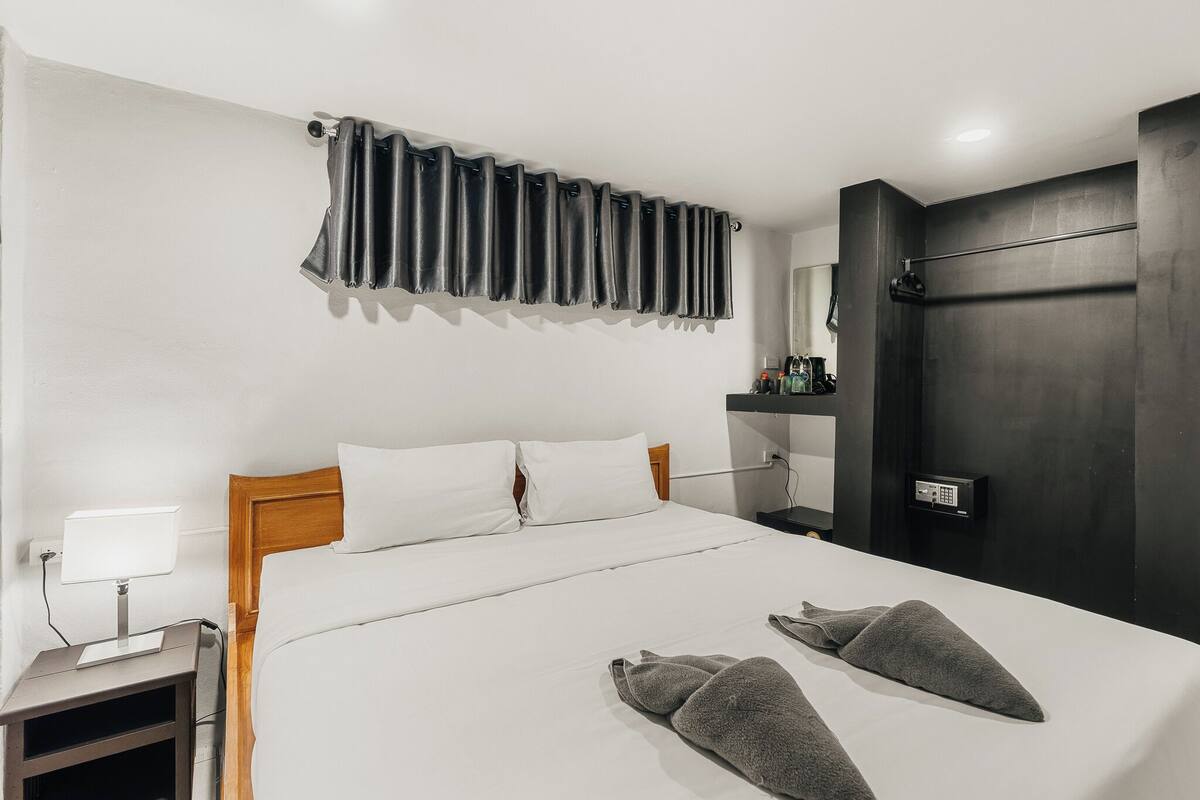 A hotel room with a bed, a nightstand with a lamp, a door, and a wall-mounted control panel.