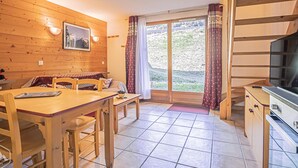 Un interior de madera con una mesa de comedor, sillas, un sofá, un televisor y un ventanal corredero que da a un balcón.