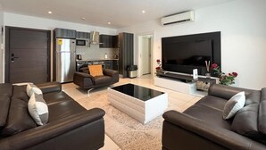 Ein modernes Wohnzimmer mit Flachbildfernseher, dunklen Sofas, einem Couchtisch und einer Küche im Hintergrund.