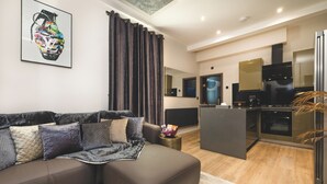 A modern living room with a sectional sofa, a small coffee table, and a kitchen area with a refrigerator, oven, and cabinets. - Private Luxury Spa Suite: Sauna, Jacuzzi, Ice Bath (Burton upon Trent)