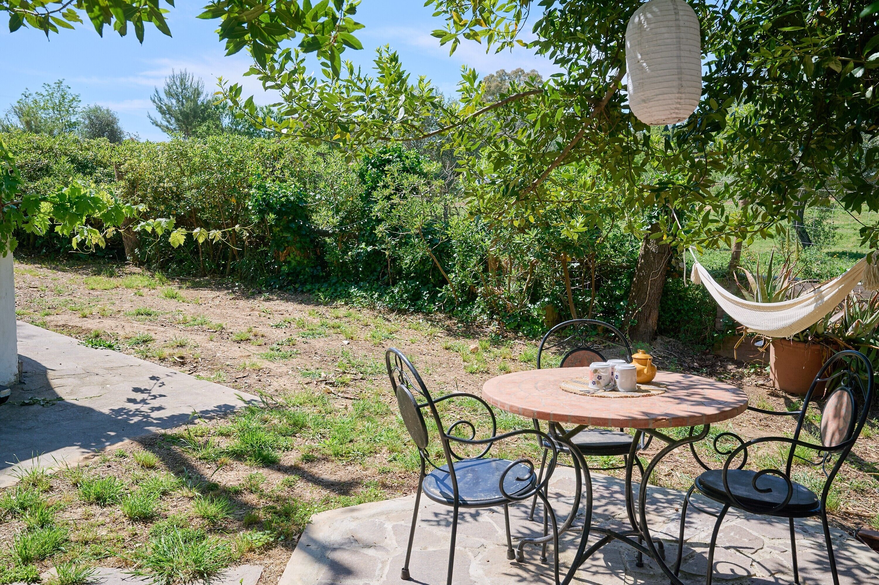 A patio with a round table and metal chairs, surrounded by greenery and a hammock.