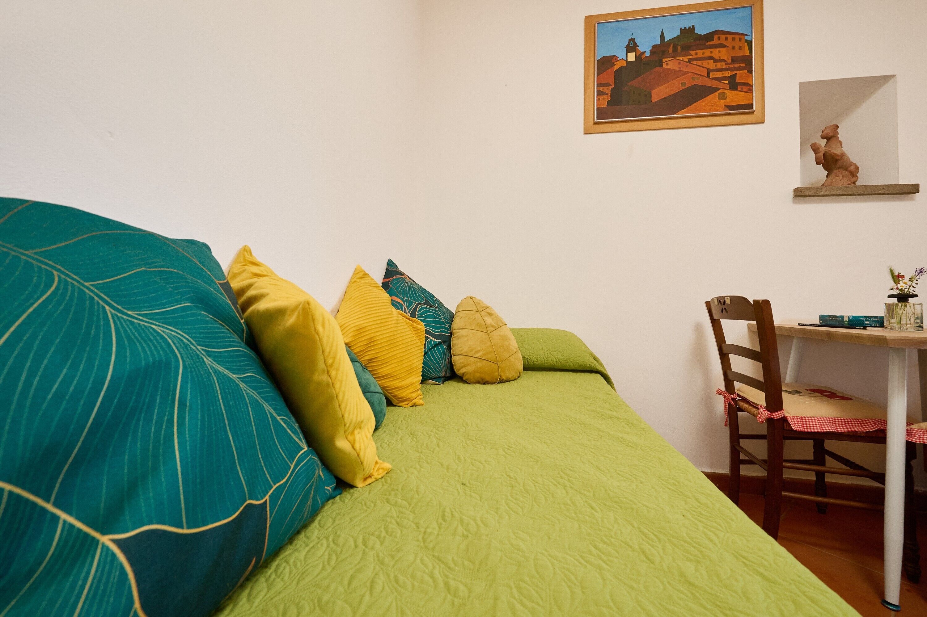 A room with a green bed, a wooden chair, a small table, and a painting on the wall.